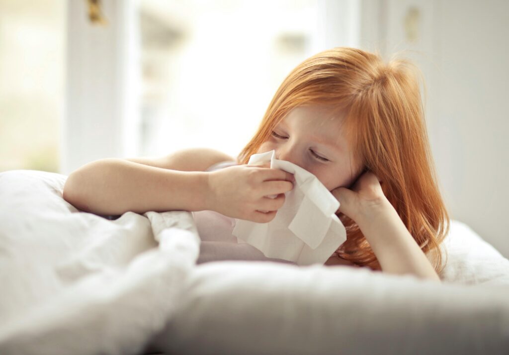 Quiet Activities for When Kids Are Feeling Under the Weather Redheaded child resting in bed, wiping nose with tissue, unwell with flu-like symptoms.