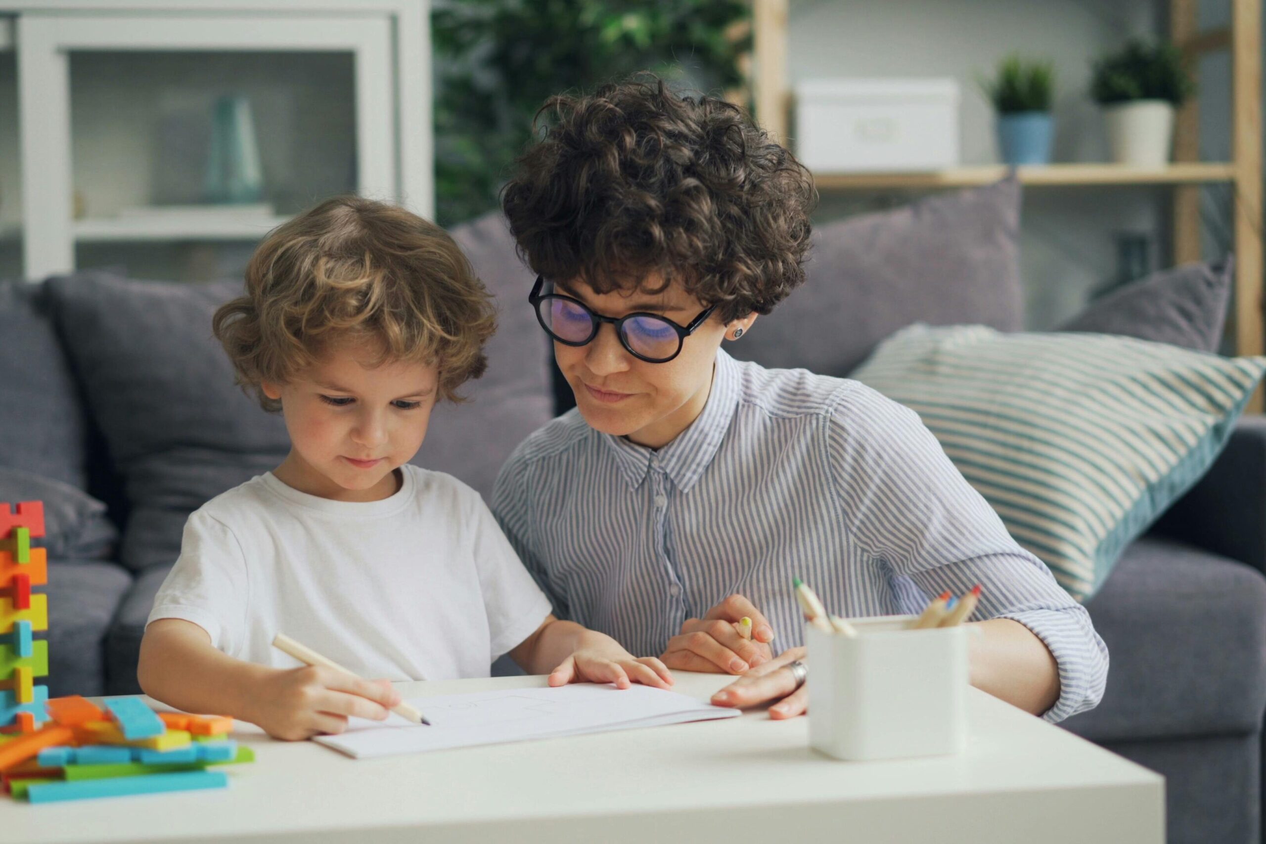 A mother helps her young son with drawing at a table in a cozy living room setting.