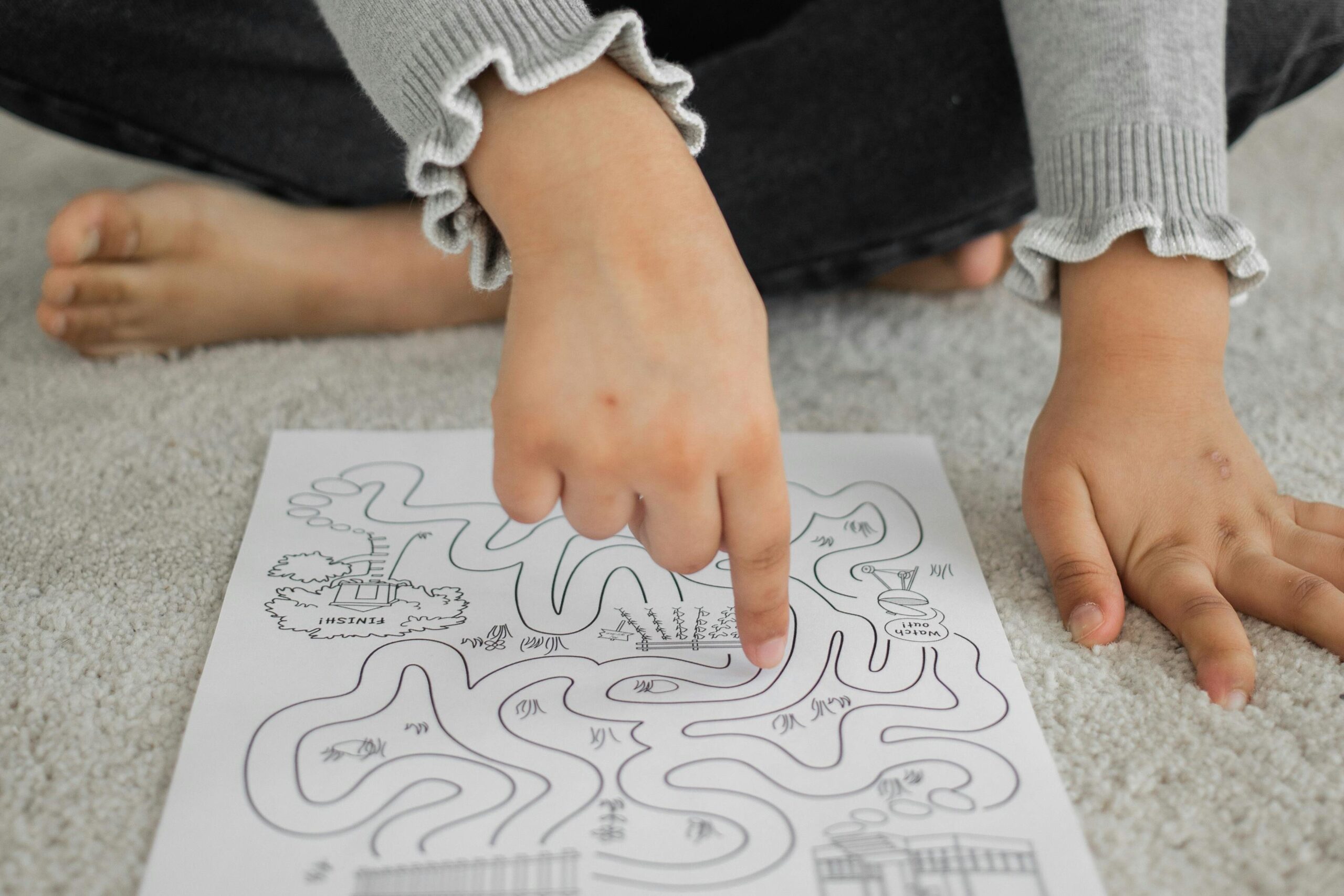 From above of crop attentive little black girl in casual clothes passing path of logic labyrinth game with finger sitting on floor at home