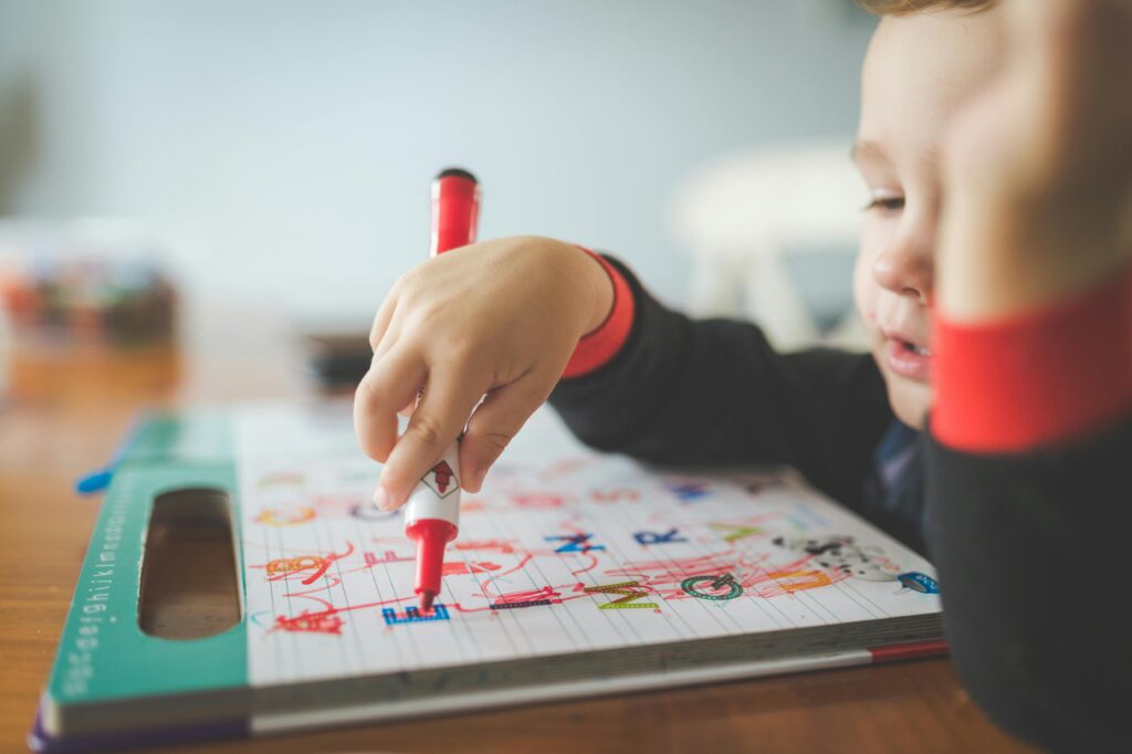A young child using a marker to write letters in an educational workbook.